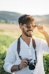 Handsome young guy in sunglasses cheerfully smiling and holding retro photo camera while standing on blurred background of amazing countryside - ADSF01029