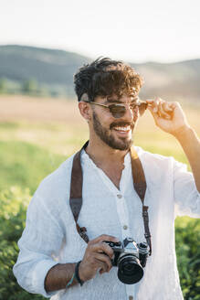 Gut aussehender junger Mann mit Sonnenbrille, der fröhlich lächelt und eine Retro-Fotokamera in der Hand hält, während er vor einem unscharfen Hintergrund mit beeindruckender Landschaft steht - ADSF01029