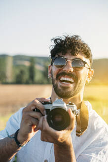 Gut aussehender junger Mann mit Sonnenbrille, der fröhlich lächelt und eine Retro-Fotokamera in der Hand hält, während er vor einem unscharfen Hintergrund mit beeindruckender Landschaft steht - ADSF01030