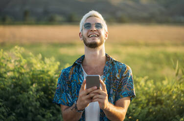 Handsome young guy in stylish outfit laughing and browsing smartphone while standing on background of beautiful nature on sunny day - ADSF01042