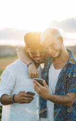 Zwei lächelnde Männer, die sich umarmen und in ihren Smartphones blättern, während sie in einer schönen Landschaft stehen - ADSF01043