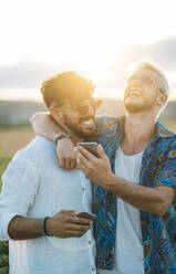 Zwei lächelnde Männer, die sich umarmen und in ihren Smartphones blättern, während sie in einer schönen Landschaft stehen - ADSF01044