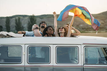 Gruppe von fröhlichen, unterschiedlichen Menschen, die in einem Retro-Van mit geöffnetem Dach stehen und eine LGBT-Flagge über den Köpfen halten, während sie gemeinsam in der Natur unterwegs sind - ADSF01047