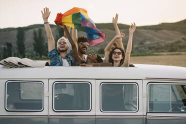 Group of cheerful diverse people standing inside retro van with opened roof and holding LGBT flag over heads while travelling in nature together - ADSF01048