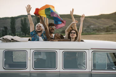 Gruppe von fröhlichen, unterschiedlichen Menschen, die in einem Retro-Van mit geöffnetem Dach stehen und eine LGBT-Flagge über den Köpfen halten, während sie gemeinsam in der Natur unterwegs sind - ADSF01048