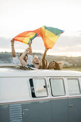 Gruppe von fröhlichen, unterschiedlichen Menschen, die in einem Retro-Van mit geöffnetem Dach stehen und eine LGBT-Flagge über den Köpfen halten, während sie gemeinsam in der Natur unterwegs sind - ADSF01049