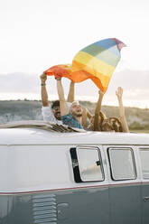 Gruppe von fröhlichen, unterschiedlichen Menschen, die in einem Retro-Van mit geöffnetem Dach stehen und eine LGBT-Flagge über den Köpfen halten, während sie gemeinsam in der Natur unterwegs sind - ADSF01050