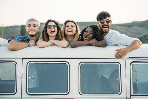 Group of diverse young friends standing in hatch in roof of vintage van and looking at camera while spending time in nature together - ADSF01054