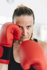 Erwachsene Frau in Boxhandschuhen, die in Kampfstellung steht und in die Kamera schaut, während des Trainings - ADSF01080