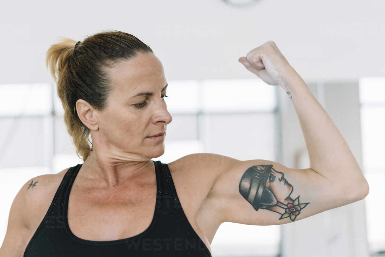 Attractive adult woman showing biceps of tattooed arm and looking at it  while training in gym stock photo