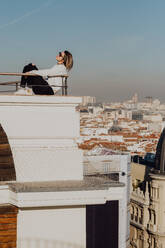 Seitenansicht der jungen stilvollen Dame in Freizeitkleidung und Sonnenbrille lehnt auf Schienen auf dem Dach und blauen Himmel auf Stadtlandschaft Hintergrund in Madrid, Spanien - ADSF01109
