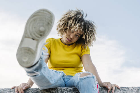 Young woman sitting on wall, showing sole of shoe - XLGF00360