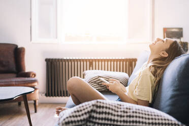 Side view of relaxed young woman sitting on couch and laughing while holding smartphone in sunlight - ADSF01162