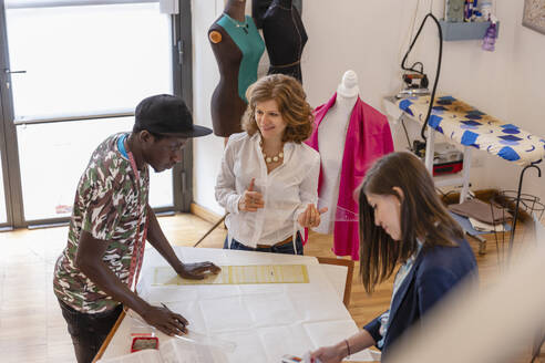 Smiling fashion designer looking at male trainee drawing at workbench at studio - EIF00084