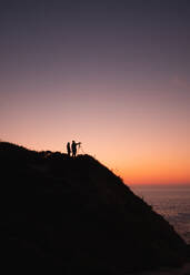 Silhouette of two people with camera on tripod standing on coast near calm sea on background of cloudless sunset sky - ADSF01172