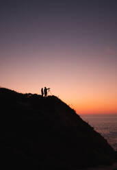 Silhouette von zwei Menschen mit Kamera auf Stativ stehen an der Küste in der Nähe von ruhigen Meer auf dem Hintergrund der wolkenlosen Sonnenuntergang Himmel - ADSF01172