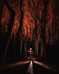 Male in hood highlighted by light and holding big wand in gloomy dark forest with red sky in Bosque Finlandes, Rascafria, Spain - ADSF01180