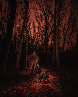 Back view human in hood holding big wand and dragging unconscious male in gloomy dark forest with red sky in Bosque Finlandes, Rascafria, Spain - ADSF01181