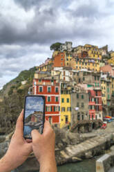 Personal perspective of man photographing Riomaggiore with smartphone, Cinque Terre, UNESCO World Heritage Site, La Spezia province, Liguria, Italy, Europe - RHPLF15446