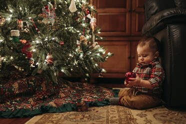 Toddler boy sitting under Christmas tree holding ornament - CAVF86951