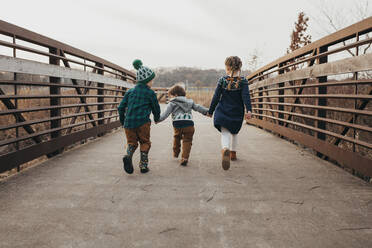 Drei Geschwister halten sich an den Händen und laufen auf einer Brücke von der Kamera weg - CAVF86956