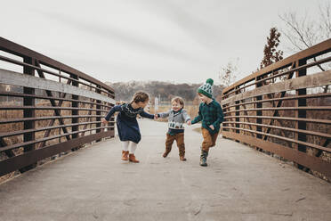 Drei lachende Geschwister, die sich an den Händen halten und auf einer Brücke laufen - CAVF86958