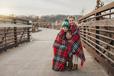 Siblings wrapped in Christmas plaid blanket on bridge at sunset - CAVF86962