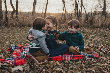 Drei Geschwister sitzen lachend auf einer rotkarierten Decke - CAVF86965