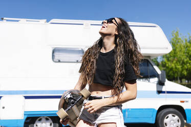 Young woman carrying skateboard in front of camper - JCMF00967