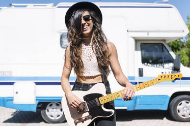 Happy young woman playing electric guitar infront of a camper - JCMF00970