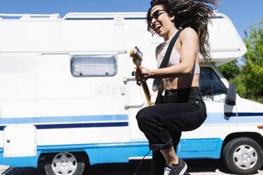 Happy young woman playing electric guitar infront of a camper - JCMF00971