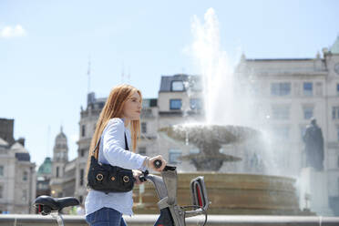 Schöne Frau zu Fuß mit Fahrrad durch Brunnen in der Stadt an einem sonnigen Tag - PMF01148