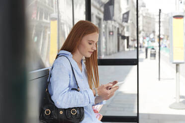 Schöne rothaarige Frau, die ein Mobiltelefon benutzt, während sie an einer Bushaltestelle in der Stadt wartet - PMF01154