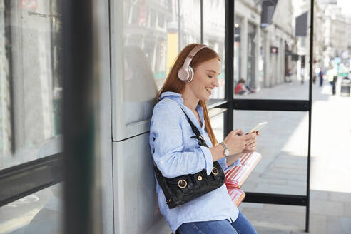 Lächelnde junge Frau hört Musik und benutzt ihr Smartphone an der Bushaltestelle - PMF01155
