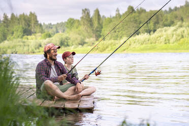 Father and son fishing while sitting on boardwalk at riverbank - VPIF02605