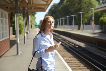 Young woman holding smart phone while standing at railroad station platform - PMF01168