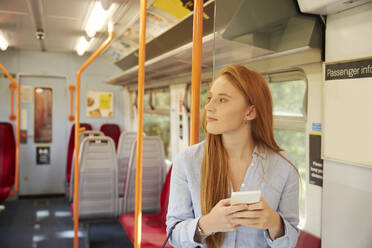 Thoughtful woman holding smart phone while standing in train - PMF01174