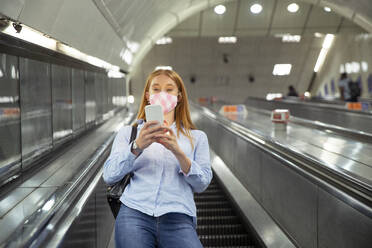 Young woman in mask using smart phone while standing on escalator at subway station - PMF01183