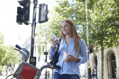 Glückliche junge Frau, die ihr Smartphone benutzt, während sie bei einem Leihfahrrad in der Stadt steht - PMF01188