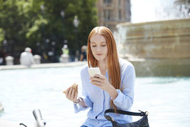 Frau, die ein Smartphone benutzt, während sie an einem Brunnen in der Stadt einen Snack zu sich nimmt - PMF01192