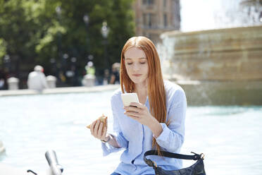 Woman using smart phone while having snacks at fountain in city - PMF01192