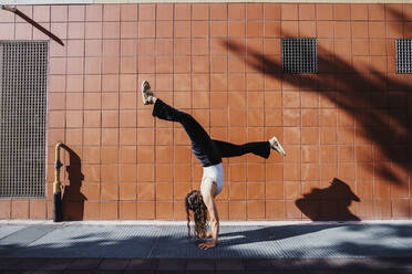 Junge Frau im Handstand auf dem Bürgersteig gegen eine geflieste Wand in der Stadt an einem sonnigen Tag - MEUF01300