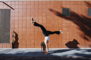 Young woman performing handstand on sidewalk against tiled wall in city during sunny day - MEUF01300