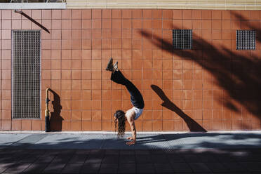 Young woman performing handstand on sidewalk against tiled wall in city - MEUF01304