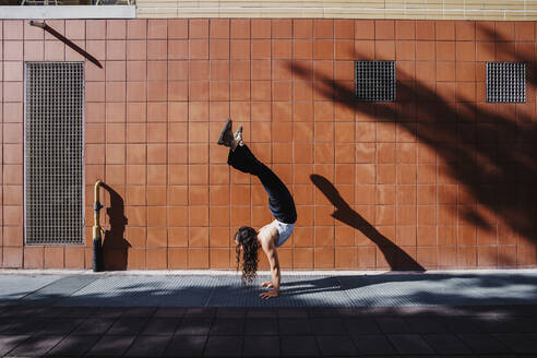 Junge Frau im Handstand auf dem Gehweg vor einer gekachelten Wand in der Stadt - MEUF01304