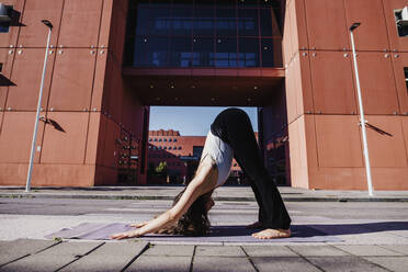Young woman performing yoga in downward facing dog position on city street - MEUF01306