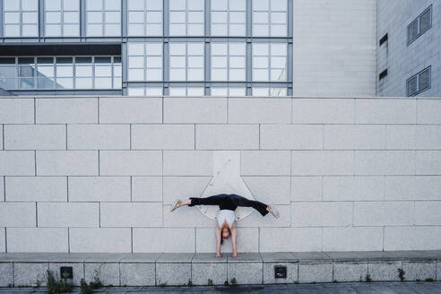 Young woman performing handstand on sidewalk against tiled wall in city - MEUF01309