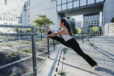 Young sporty woman stretching legs on railing in city during sunny day - MEUF01312