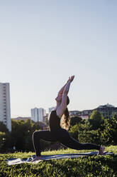 Young woman practicing yoga in warrior position at city park against clear sky - MEUF01324