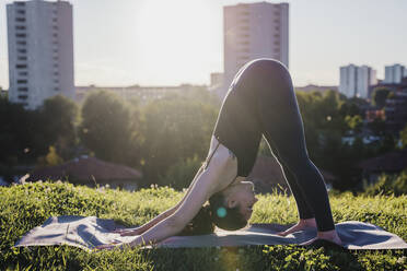 Young woman performing yoga in downward facing dog position at city park - MEUF01325